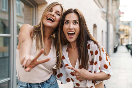 Image of excited adult two women hugging and gesturing peace sign while walking on city street