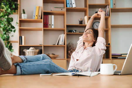 Image of pleased adult woman stretching her body while working and sitting with legs on table in...