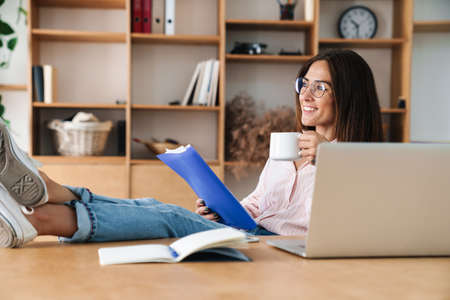 Image of smiling businesswoman drinking coffee and reading documents while sitting with legs on...