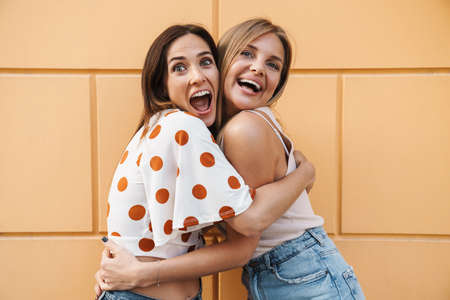 Image of excited adult two women hugging and laughing while posing on city street