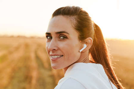 Image of happy brunette sportswoman using earphones and smiling while working out at golden field