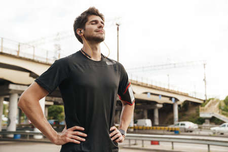 Image of athletic pleased sportsman with smartphone using earphones while working out on city street