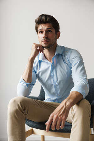Image of serious businesslike brunette man thinking and sitting on chair over white wall in room