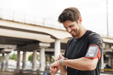 Image of athletic smiling sportsman with smartphone using smartwatch while working out on city...