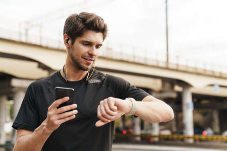 Image of young sportsman in earphones using cellphone and smartwatch while working out on city streetの写真素材