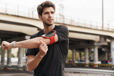 Image of athletic young sportsman with smartphone doing exercise while working out on city street