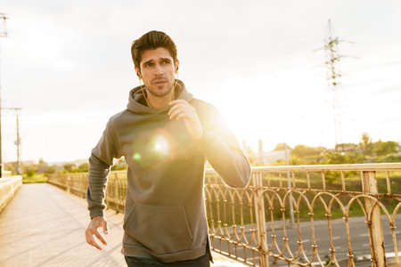 Image of athletic caucasian sportsman using earphones while running on city bridge