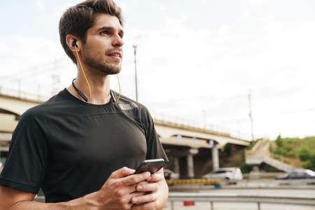 Image of handsome young sportsman in earphones using cellphone while working out on city streetの写真素材