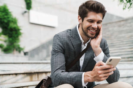 Image of smiling positive handsome business man outdoors using mobile phone while sitting on the stairsの写真素材