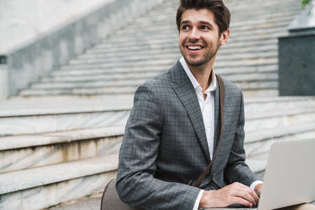 Image of cheerful handsome business man outdoors using laptop computer while sitting on the stairsの写真素材