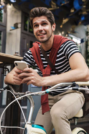 Image of cheerful handsome man smiling and using cellphone on bicycle at city streetの写真素材