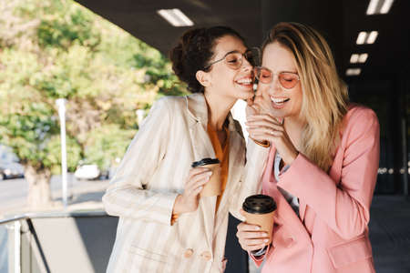 Two beautiful smiling young businesswomen walking outdoors at the city streets, talking while...