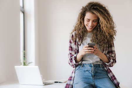 Photo of cheerful beautiful woman smiling and using mobile phone while sitting on table in white...
