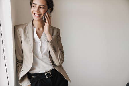 Photo of cheerful successful woman in jacket talking on mobile phone while leaning on wall at officeの写真素材