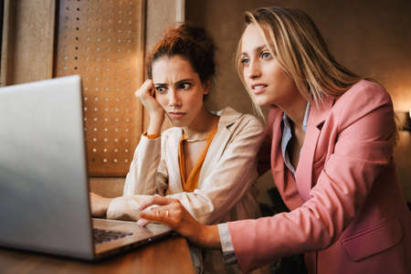 Image of a shocked emotional two business women colleagues indoors in cafe using laptop computer.の写真素材