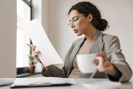 Photo of focused brunette woman in eyeglasses drinking coffee while working with documents in white...