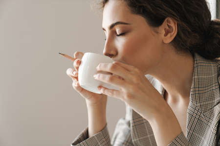 Photo closeup of pleased brunette woman drinking coffee while working in white officeの写真素材