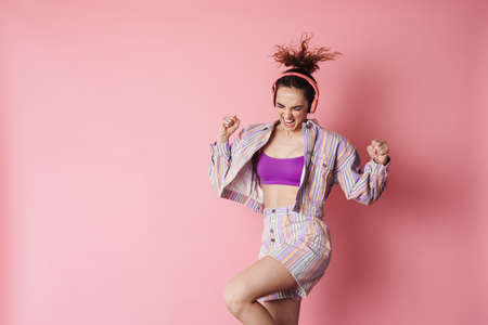 Image of ginger happy woman dancing while using wireless headphones isolated over pink backgroundの写真素材