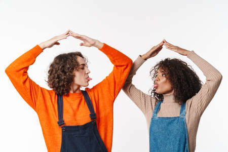 Portrait of funny multinational women in overalls making roof above their heads with arms isolated...