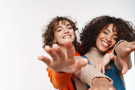 Image of a beautiful happy cheery optimistic two multiracial girls friends in denim jumpsuits isolated over white wall background having fun raises hands to camera.の写真素材