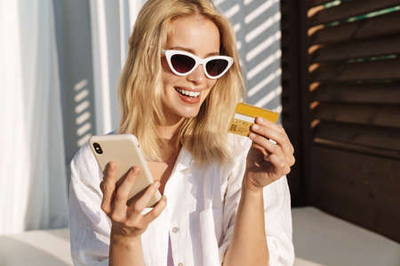 Image of excited woman in sunglasses holding credit card and using cellphone at hotel on summer...