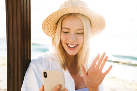 Image of cheerful blonde woman using mobile phone while standing at summer beachの写真素材