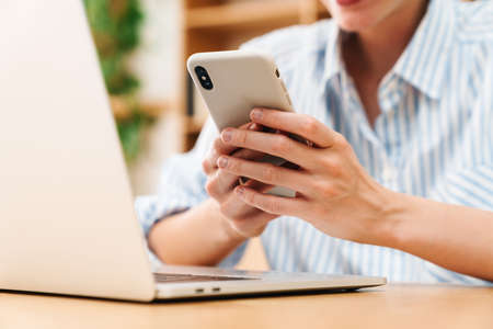 Cropped image of woman typing on mobile phone while working with laptop at table in office