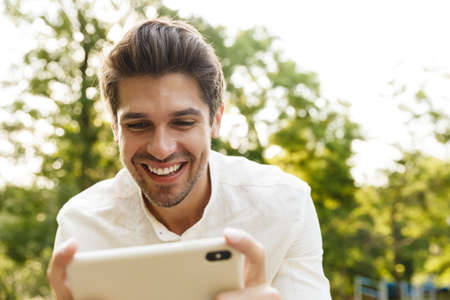 Image of young caucasian brunette man smiling and holding cellphone while walking in parkの写真素材