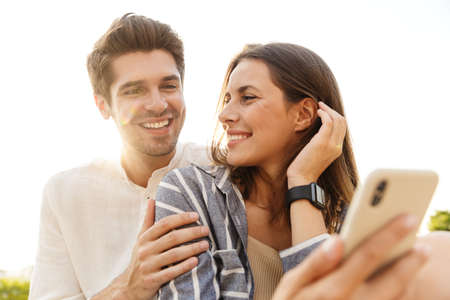 Image of young caucasian happy couple smiling and hugging while sitting outdoors with cellphone