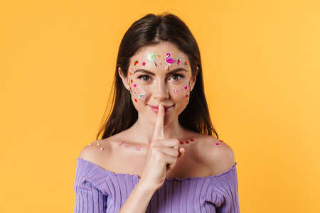 Image of cheerful woman with stickers on face showing silence gesture isolated over yellow...