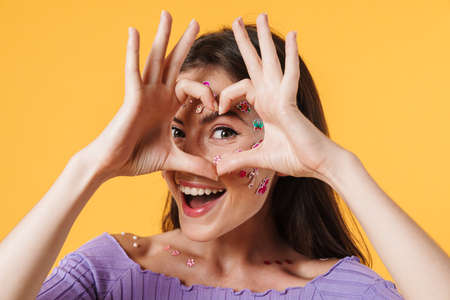 Image of young joyful woman with stickers on face showing heart gesture isolated over yellow backgroundの写真素材