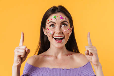 Image of smiling woman with stickers on face pointing fingers upward isolated over yellow wallの写真素材
