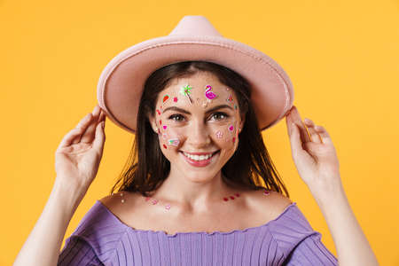 Image of young cheerful woman with stickers on face posing in pink hat isolated over yellow...