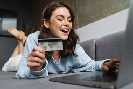 Image of cheerful nice woman holding credit card and working with laptop while lying on sofa at home