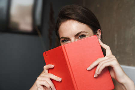 Image of beautiful joyful woman putting diary over her face and looking at camera at homeの写真素材
