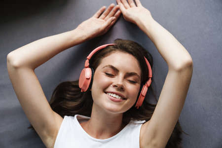 Image of beautiful joyful woman smiling and using wireless headphones while lying at home