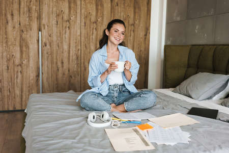 Image of a young happy cheery girl indoors at home drinking coffee while sitting near paper...