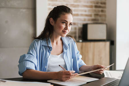 Photo of a young cute positive girl indoors at home using laptop computer and studying