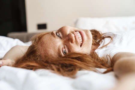 Image of ginger happy woman with freckles smiling at camera while lying in bed at homeの写真素材