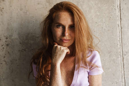 Image of redhead sensual woman looking at camera while posing near concrete wall at homeの写真素材