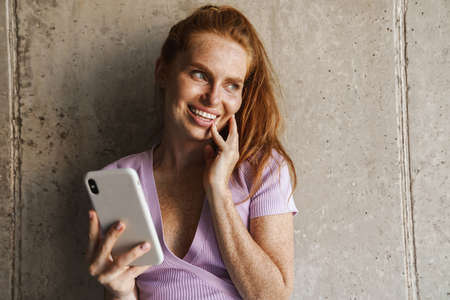 Image of redhead joyful woman smiling and using cellphone while standing near concrete wall at homeの写真素材