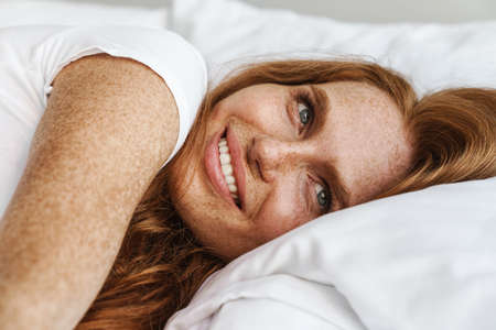 Image of ginger happy woman with freckles smiling at camera while lying in bed at homeの写真素材