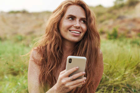 Happy young woman in summer dress sitting in park, texting on mobile phoneの写真素材