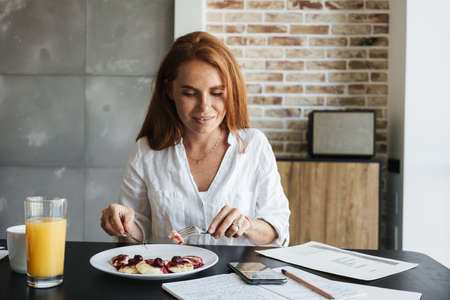 Image of pleased ginger businesswoman using cellphone while having breakfast in kitchen at home