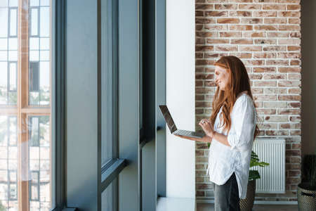 Image of redhead smiling businesswoman making video call on laptop while standing near window in homeの写真素材