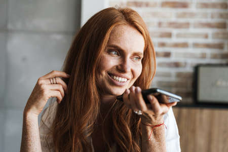 Image of happy ginger woman with freckles smiling and talking on cellphone in homeの写真素材