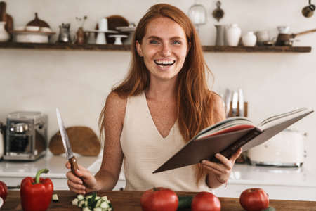 Image of happy ginger woman taking reading cooking book while preparing lunch at cozy kitchenの写真素材