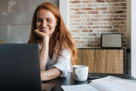 Image of smiling redhead businesswoman working with laptop while sitting at table in homeの写真素材