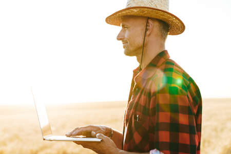 Image of pleased adult man in straw hat working with laptop while standing at cereal fieldの写真素材
