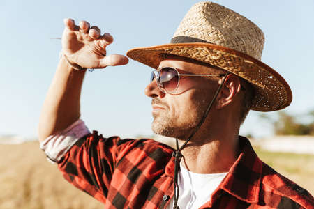 Image of unshaven adult man in straw hat and sunglasses looking aside while standing at cereal fieldの写真素材